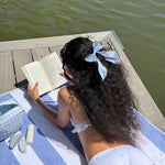 Bow Barrette in Baby Blue Gingham model reading a book while wearing Bow Barrette in Baby Blue Gingham in hair and laying on striped towel across wooden dock next to emi jay hair care and accessories