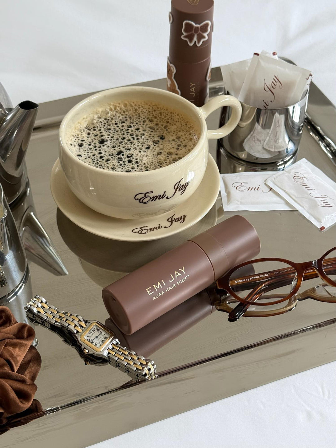 Aura Hair + Body Mist in Toasted sugar on tray with Emi Jay coffee cup, Emi Jay sugar packets, watch, scrunchie, and glasses. Aura Hair + Body Mist in Toasted sugar on tray with Emi Jay coffee cup, Emi Jay sugar packets, watch, scrunchie, and glasses.