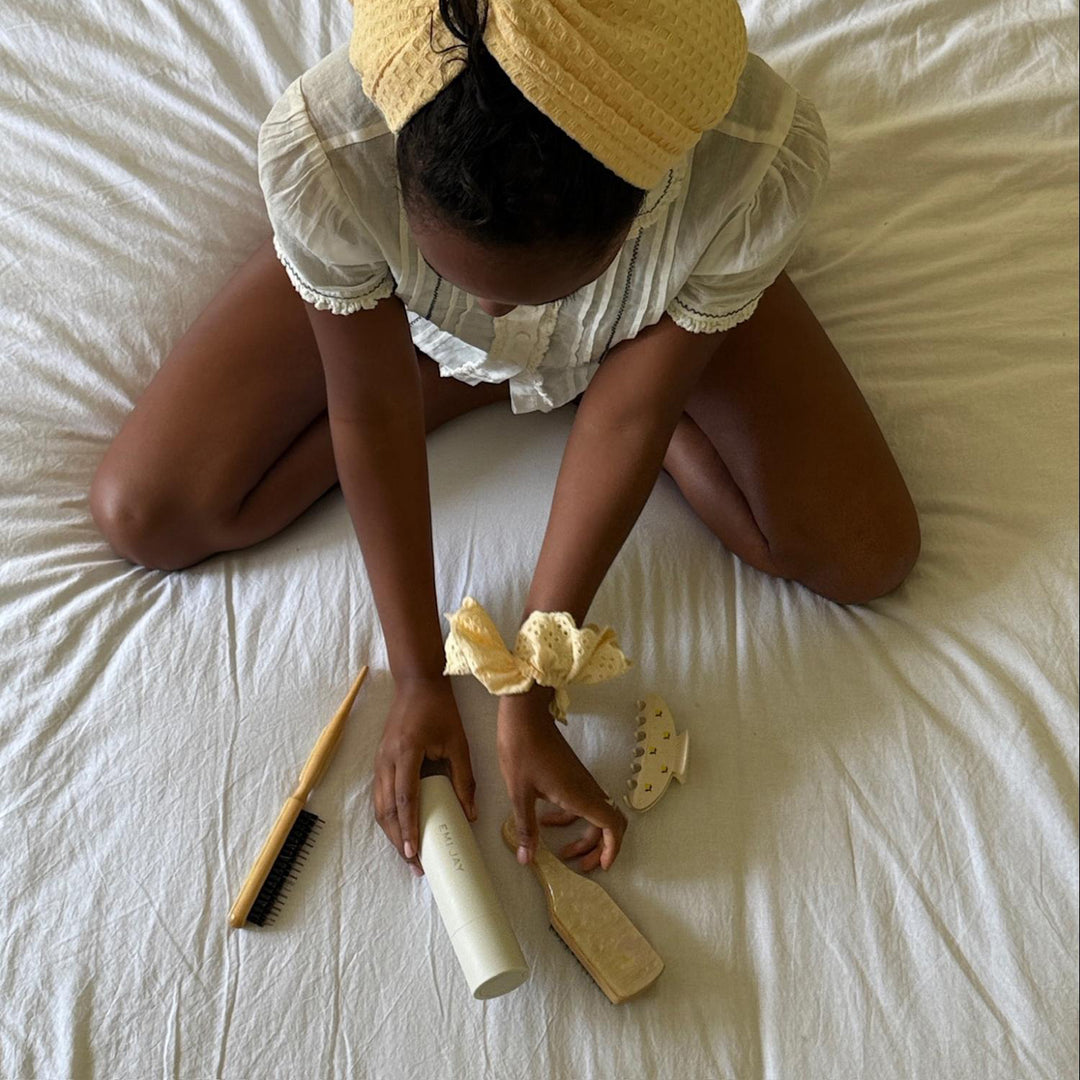 model with hair in towe and eyelet scrunchie on wrist sitting next to styling brush in lemon sugar and big effing clip while picking up heavenly hair milk and mini boar bristle brush