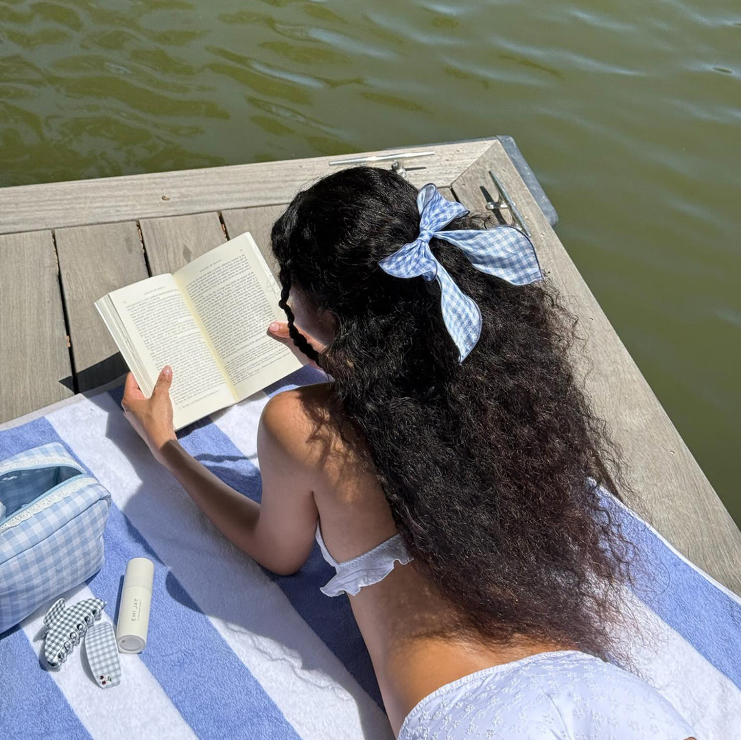 model reading a book while wearing Bow Barrette in Baby Blue Gingham in hair and laying on striped towel across wooden dock next to emi jay hair care and accessories 