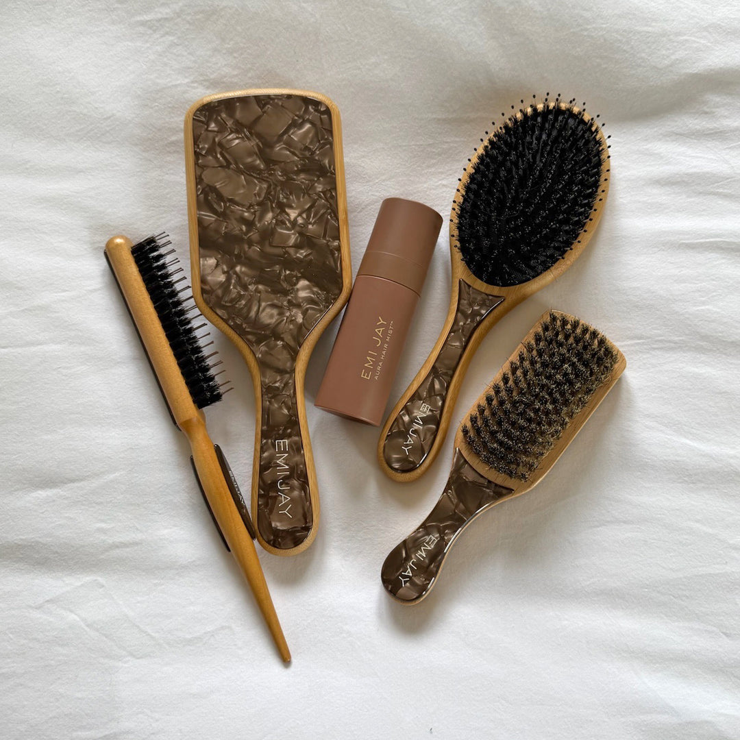 Assorted Emi Jay hair brushes with Aura Hair + Body Mist in Toasted Sugar on bed.