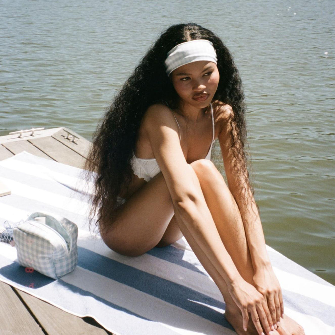 model sitting on wooden dock next to pouch while wearing Infinity Headband in Baby Blue Gingham in hair