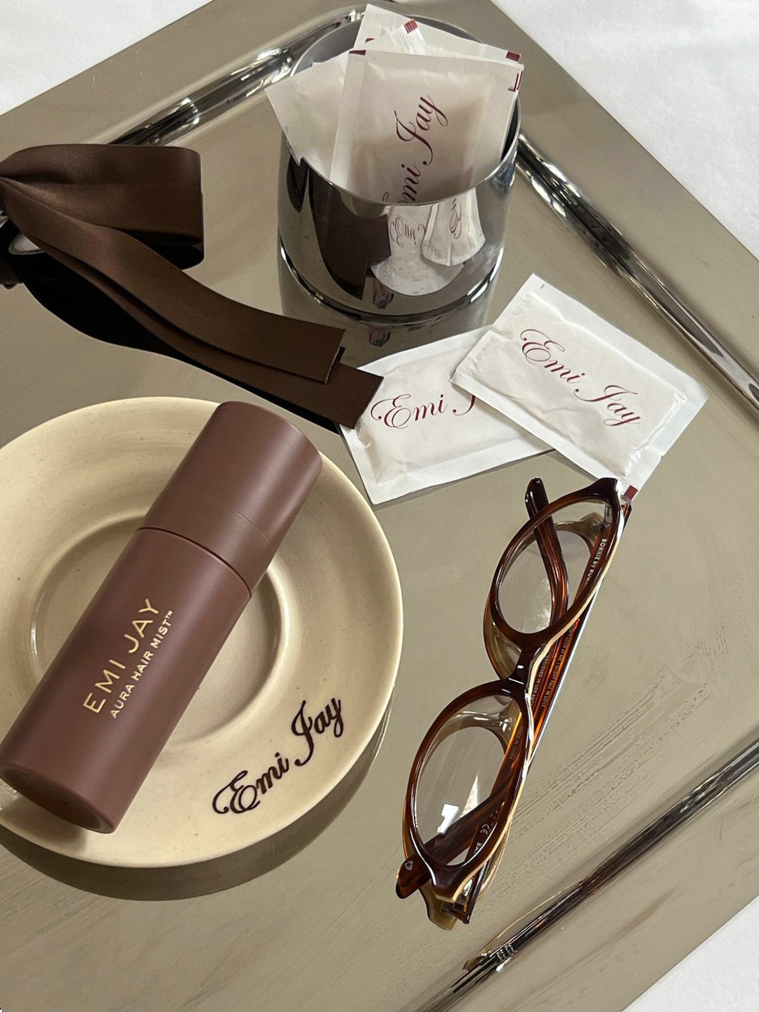 Aura Hair + Body Mist in Toasted sugar on tray with Emi Jay coffee cup, Emi Jay sugar packets, watch, scrunchie, and glasses.