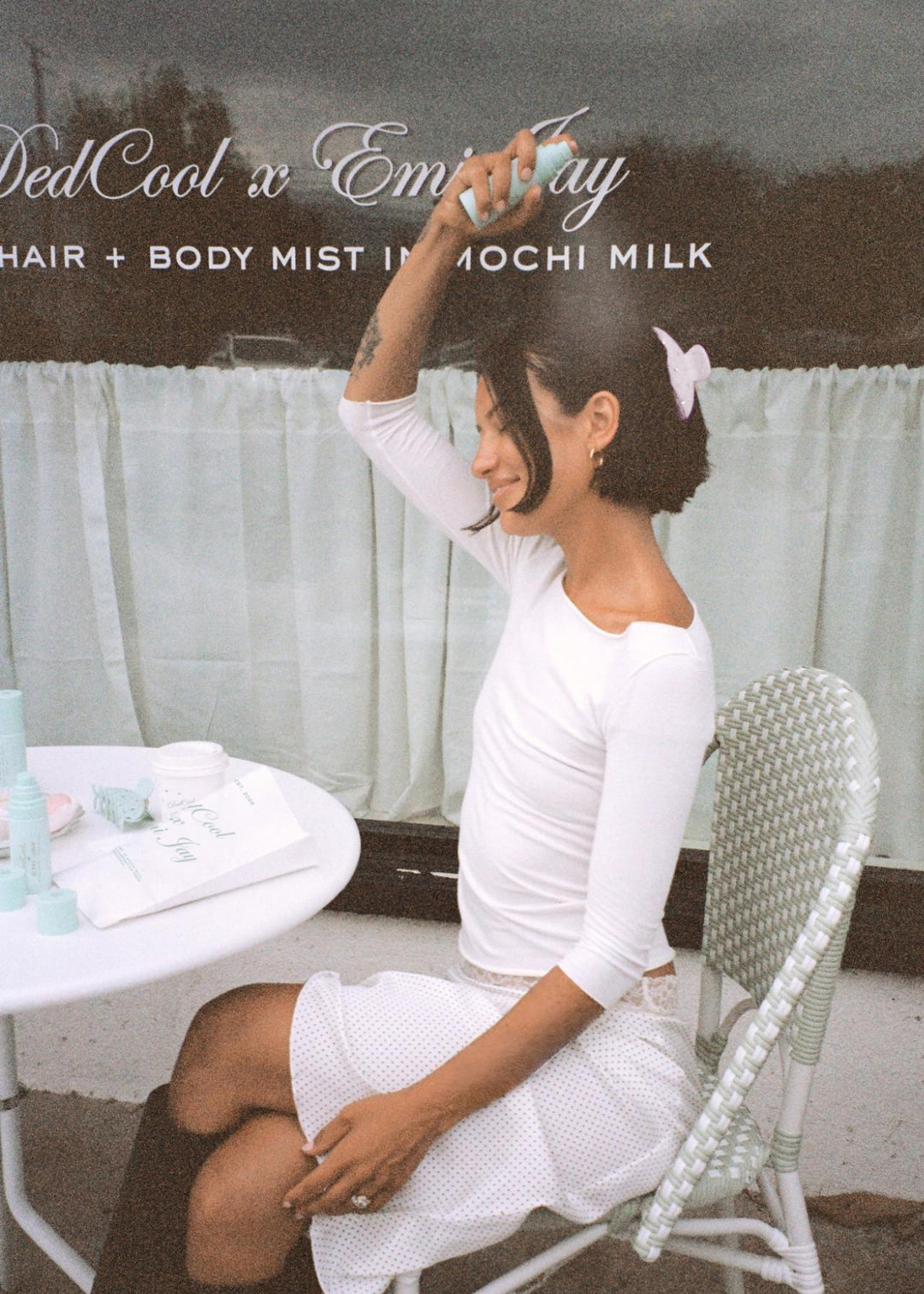 model spraying aura hair + body mist in mochi milk in hair while wearing big effing clip and sitting at table in front of DedCool x Emi Jay window
