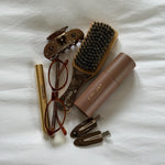 Assorted Emi Jay hair care, accessories, and styling tools with glasses on bed.