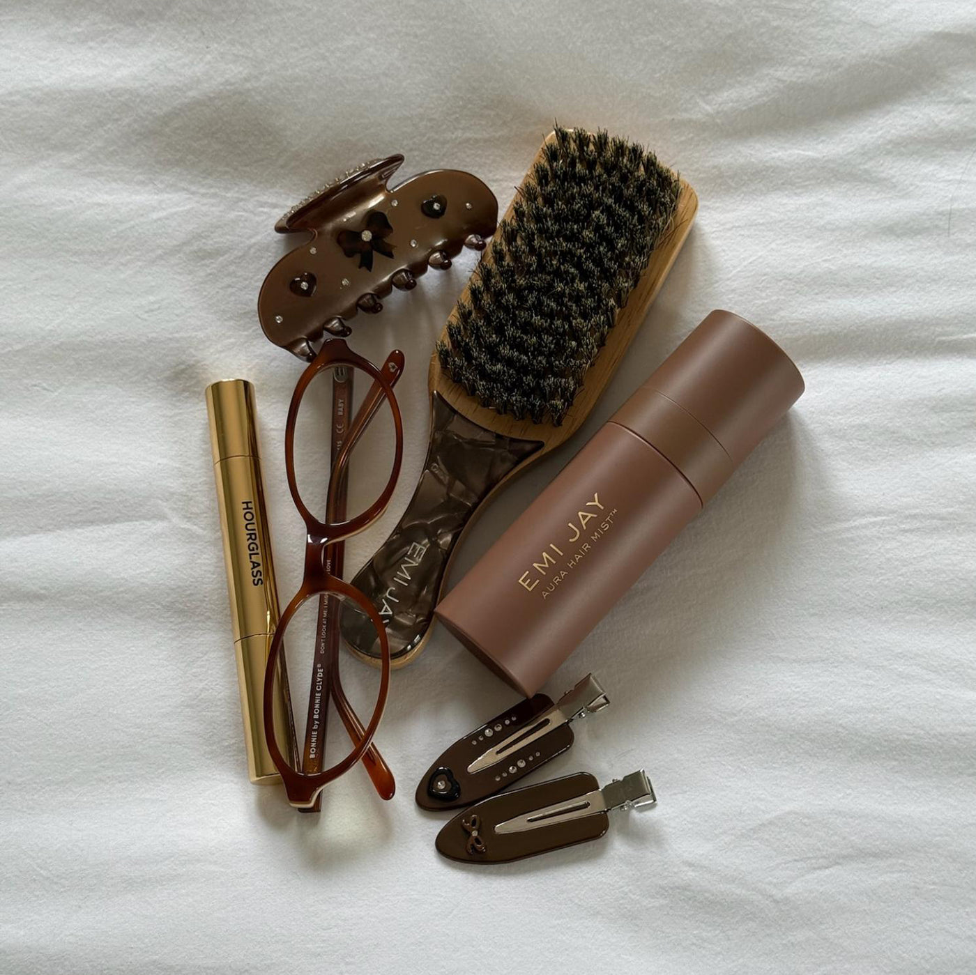 Assorted Emi Jay hair care, accessories, and styling tools with glasses on bed.
