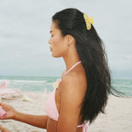 model standing on beach holding aura hair + body mist in lychee baby while wearing Sweetheart Clip in Citrus Baby Bloom in hair
