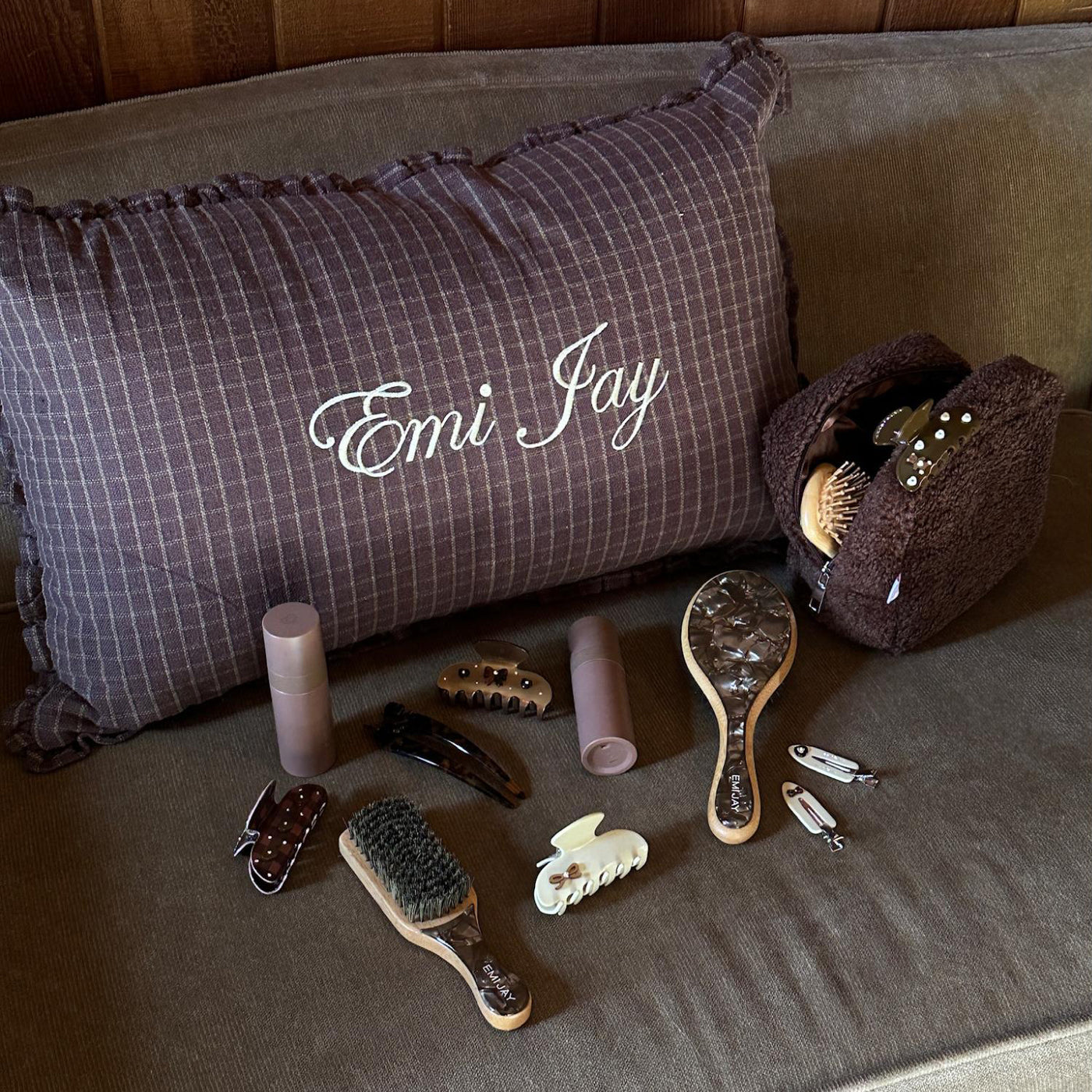 Assorted hair care, accessories, and pouch next to Emi Jay embroidered pillow on couch.